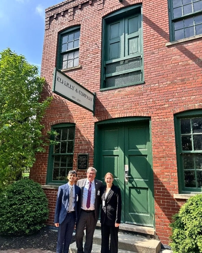 Three people in business attire standing outside a brick Eli Lilly building on a sunny day.