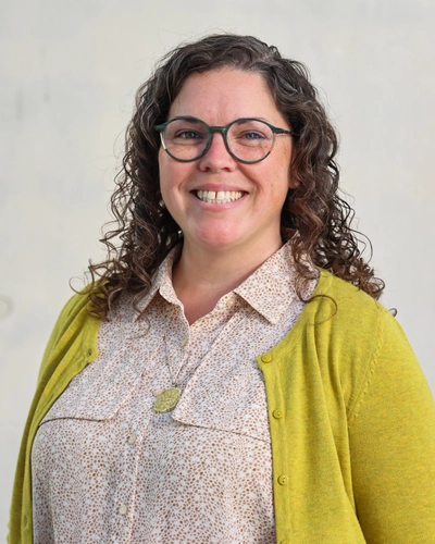 A woman with curly brown hair and glasses smiles while standing against a light gray background.
