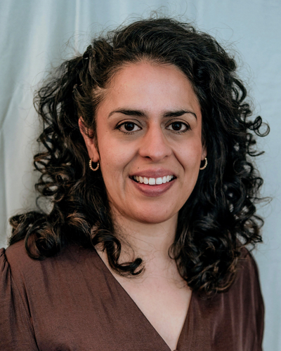 Close-up portrait of a person with dark, curly hair wearing small hoop earrings and a brown top, facing the camera against a soft, light-colored background.
