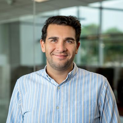 Person wearing a light blue and white vertically striped button-up shirt, standing indoors in front of large glass windows with a blurred office background.