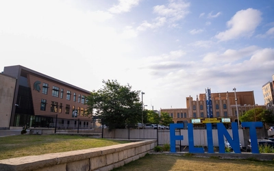 Large blue letters spelling 'FLINT' in front of downtown Flint, Michigan. Behind the sign is the Michigan State University College of Human Medicine public health building with a Spartan helmet logo, and the historic Capitol Theatre is visible in the background.