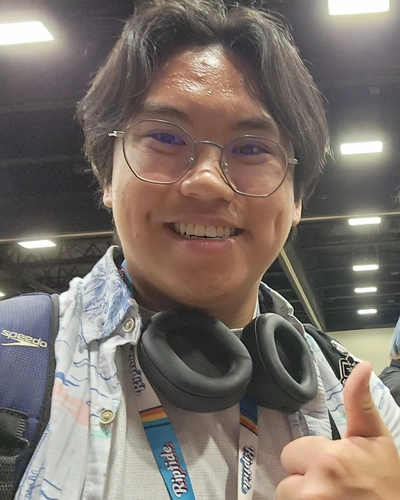 A selfie of a person smiling and giving a thumbs up while wearing a backpack, headphones around their neck and glasses.