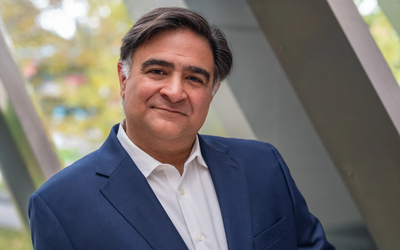 Person wearing a dark blue suit jacket over a white dress shirt, standing outdoors in front of large angled metal beams with blurred greenery in the background