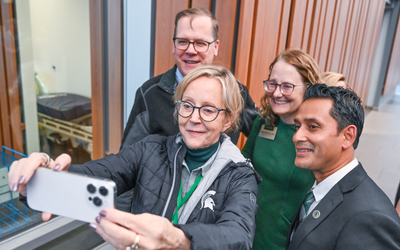 A group of people stand near a wall with vertical wooden panels, posing for a selfie. One person in the foreground holds a smartphone at arm’s length to capture the photo. Visible behind the group is a large window with a view of a room containing a bed or examination table.