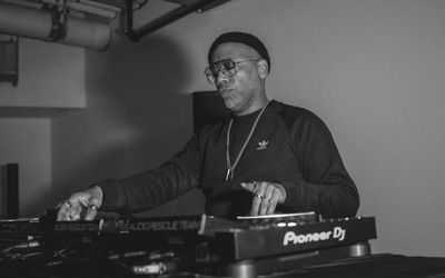 A DJ performs in a dimly lit room, standing behind a set of Pioneer turntables and audio equipment. They wear a dark long‑sleeve shirt with a small logo and a chain necklace, with one hand adjusting controls on the mixer. The scene is captured in black and white, highlighting the industrial ceiling pipes above and the focused atmosphere of the performance.