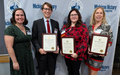 Four individuals standing on a wooden floor in front of a backdrop featuring the Michigan History Magazine and Historical Society of Michigan logos. Three of them are holding framed award plaques with visible text and gold seals at the bottom. All are dressed in formal or semi-formal attire, and each has a name badge pinned to their clothing.