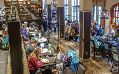 Students eat in a dining hall with ornate lighting and a bookshelf in the background
