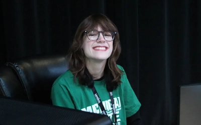A person wearing glasses with long, reddish hair smiles as she sits in front of a computer screen