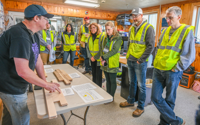 Person wearing a black T-shirt stands at a table indoors, demonstrating wooden pieces and printed instructions to a group of people. The group is wearing bright yellow-green safety vests and casual clothing, gathered closely around the table. The room has wood-paneled walls, windows, and shelves with various items in the background