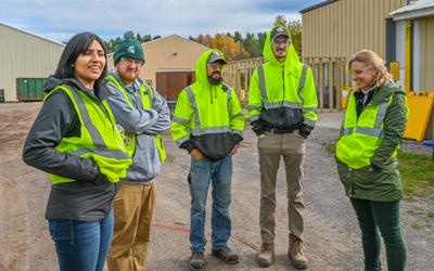 Five people wearing bright yellow-green safety vests and outdoor clothing stand in a group on a gravel surface between large beige industrial buildings. Some individuals have their hands in their pockets, and two are wearing green caps. The background includes storage containers, fencing, and trees with autumn foliage.