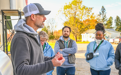 Person wearing a gray jacket and white mesh cap speaks to a small group outdoors near a building. The group members are dressed in jackets and have green lanyards with name badges. Behind them are trees with autumn foliage, a gravel area, and a white house in the background