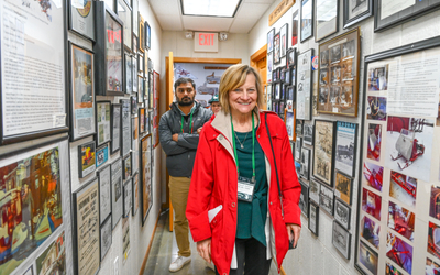 “Person wearing a bright red coat and green lanyard walks through a narrow hallway lined with framed photographs, articles, and memorabilia on both walls. Two other individuals follow behind, also wearing lanyards. The hallway has a white ceiling with fluorescent lights and an exit sign above a doorway at the end