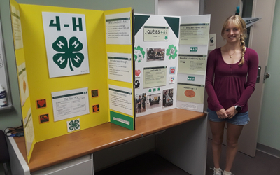 A tri-fold presentation board about 4‑H is displayed on a desk in an office or classroom, featuring yellow and white panels with text, images, and a prominent green four-leaf clover emblem. The board includes sections such as “The Four H’s” and “Who is involved in 4‑H?”, along with additional information in Spanish. A person stands beside the display near a door, with office furniture visible nearby, suggesting an educational presentation setting.