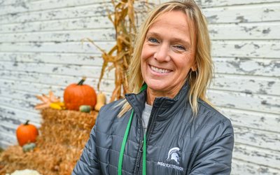 Person wearing a dark gray MSU Extension jacket with a green lanyard stands in front of a rustic white wall. Behind them is a fall-themed display featuring stacked hay bales, orange pumpkins, corn stalks, and small decorative gourds