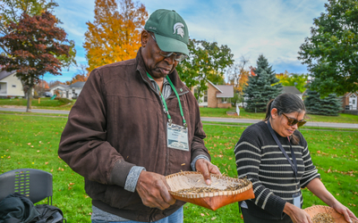 Two people stand outdoors on a grassy area with autumn trees and houses in the background. Both are holding wooden trays filled with seeds or grains, appearing to demonstrate or sort them. One person wears a dark jacket and green cap with a Spartan logo, while the other wears a striped sweater. Nearby is a black chair and a bag on the ground.
