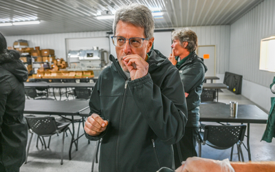 Person wearing a dark jacket stands indoors in a room with gray metal walls and ceiling, holding a small item near their mouth. The background shows black tables and chairs arranged in rows, shelves with boxes, and industrial equipment. Another person in similar attire is visible behind them, and a hand in the foreground holds a small container with a yellow substance