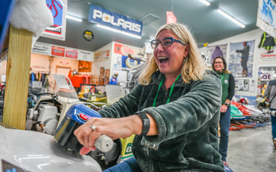 A person sits on a vintage snowmobile inside a museum filled with snowmobiles and related memorabilia. The individual grips the handlebars of a snowmobile arcade game, which has a white front panel and a blue control area. The background features multiple colorful snowmobiles, posters, and banners, including a prominent “Polaris” sign hanging from the ceiling. The setting is well-lit with fluorescent lights, and the floor is concrete.