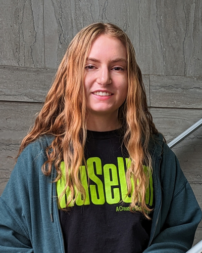 Person with long, wavy, light brown hair wearing a dark T-shirt with bright green text that reads “MUSEUM” and a tagline underneath, partially visible. Over the T-shirt, the person has on a teal zip-up jacket. The background features a gray stone wall with horizontal lines and a metal handrail on the right side, suggesting an indoor stairway or architectural setting.