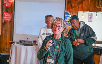 A person speaks into a microphone while standing indoors in front of a projector screen. The individual is wearing a green jacket with a lanyard and name badge. Behind the speaker, two other people are visible, one wearing a Michigan State sweatshirt and another in a dark jacket. The setting includes wooden walls, string lights, and heart-shaped wall decorations, creating a rustic atmosphere. A small round table with a projector and cup is positioned near the screen.