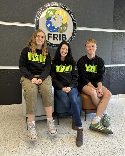 Three people are seated on small, modern stools in front of a dark wall featuring the FRIB logo, which stands for Facility for Rare Isotope Beams. The logo includes a circular design with scientific symbols and text around the edge. All three individuals are wearing matching black sweatshirts with bright green text that reads “Museum” across the front. The person on the left is wearing light-colored cargo pants and sneakers, the person in the middle is wearing blue jeans and brown boots, and the person on the right is wearing shorts with green-and-white sneakers.