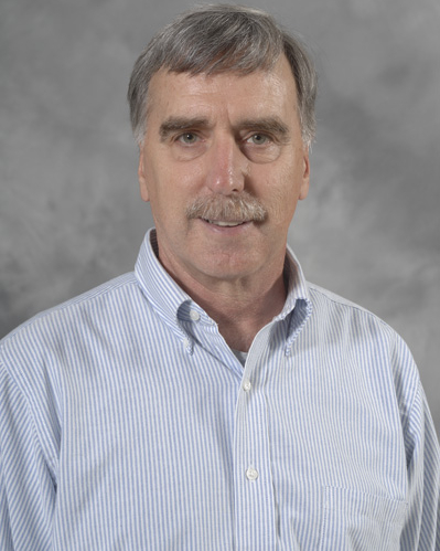A person wearing a light blue and white striped button-down shirt stands in front of a plain gray studio backdrop.
