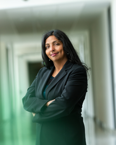 Person wearing a black blazer and dark top, standing with arms crossed in a bright hallway with glass walls and a soft green reflection in the foreground.