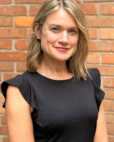 A woman wearing a short-sleeved black dress with ruffled shoulder details, standing in front of a brick wall. Her hair is shoulder-length and styled in loose waves with a middle part.