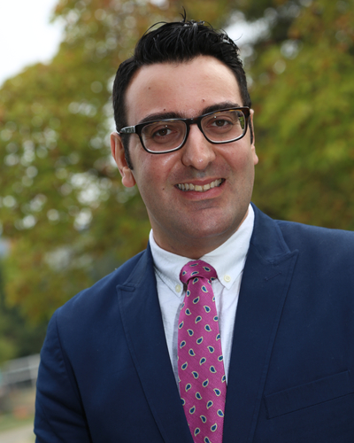 Head-and-shoulders portrait of a man wearing glasses, a navy suit, white shirt, and pink patterned tie, smiling outdoors with fall foliage in the background.