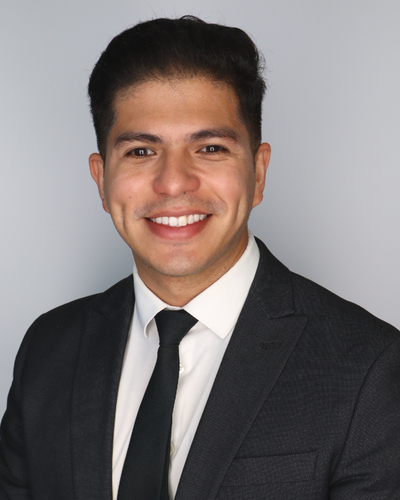 Studio head-and-shoulders portrait of a person wearing a dark suit jacket, white shirt, and black tie against a light gray background.