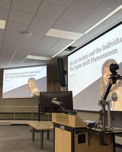 A classroom at Michigan State University shows a lecture slide for a social science course titled 'ISS 210: Society and the Individual – The Taylor Swift Phenomenon.' The slide features an image of Taylor Swift smiling.