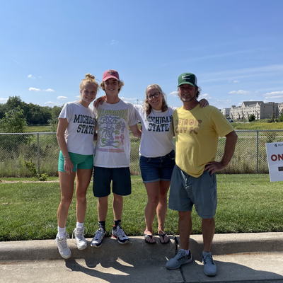 Four people pose for a photo in a parking lot. Their arms are linked. Behind them is a grass field and a building.