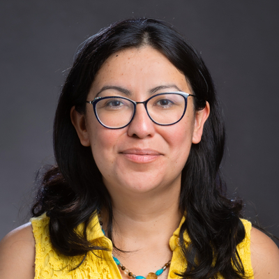 A person with shoulder‑length dark hair wearing a sleeveless yellow top and a beaded necklace, posed against a dark studio background.