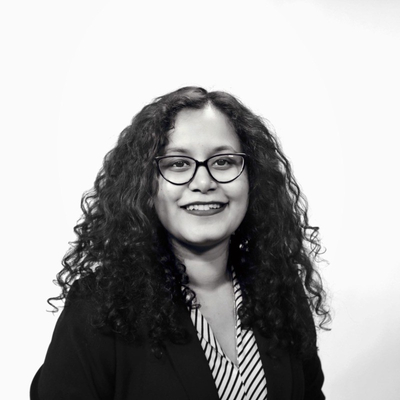 Black-and-white studio portrait of a person with curly hair and glasses, wearing a striped blouse and dark blazer.
