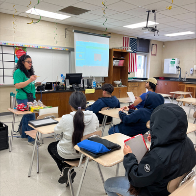 Instructor speaking at the front of a classroom while students sit at desks holding papers and looking toward a projected screen.