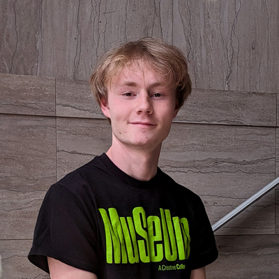 A person stands indoors in front of a wall with a stone or tile pattern, wearing a black t-shirt with bright green stylized text that reads "MuSeUm." The M, S and U are capitalized. A metal railing is visible in the background, suggesting the setting may be a public or institutional space like a museum or gallery.