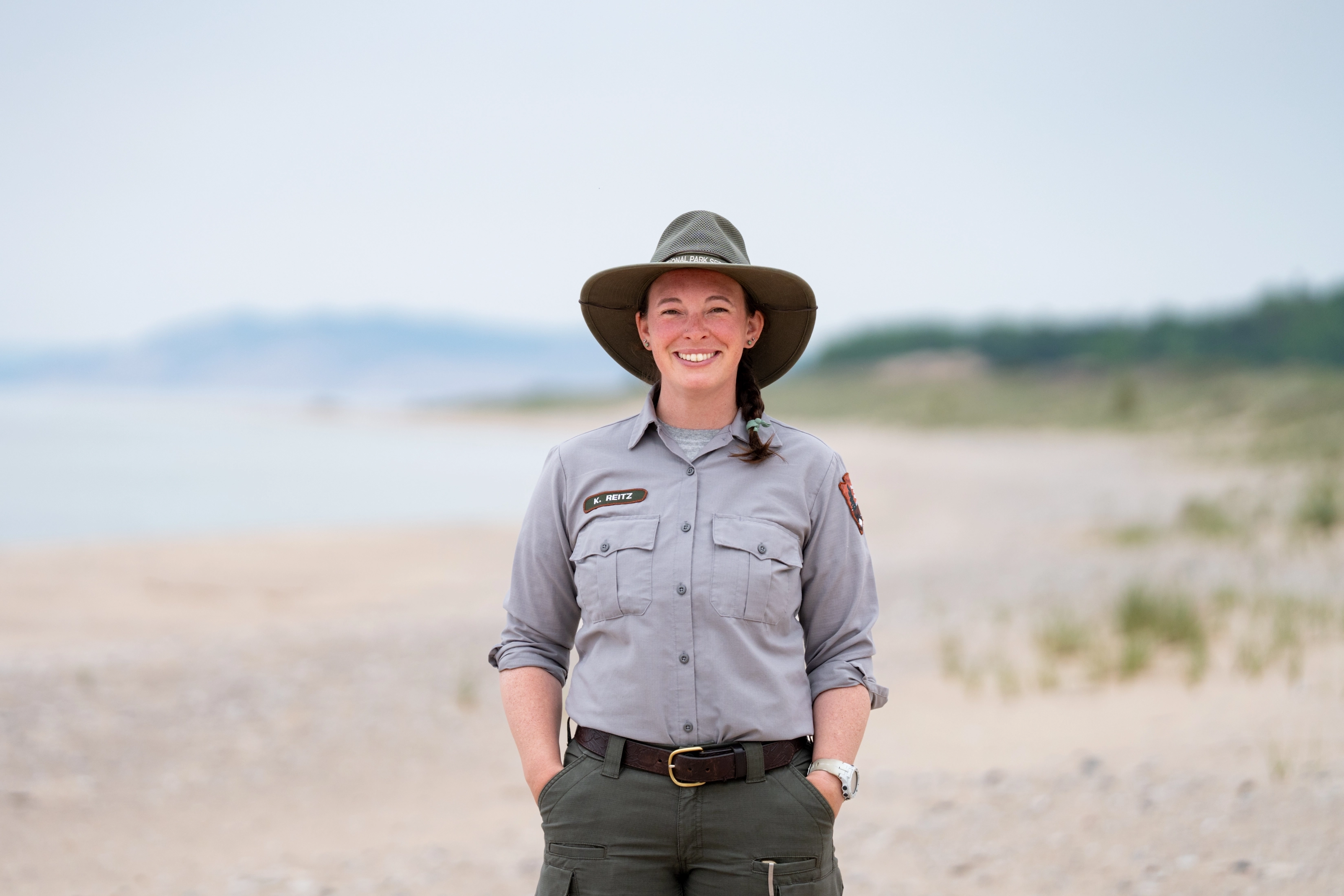 A smiling National Park Service ranger in uniform and sun hat stands on the beach at Sleeping Bear Dunes.