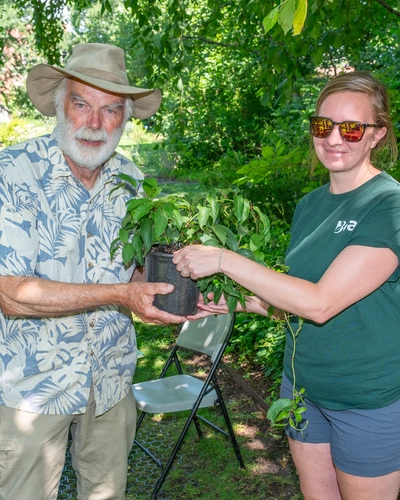 Two people standing outdoors in a garden, holding a potted plant together. One wears a hat and patterned shirt, the other a green t-shirt and shorts. Trees, greenery, and an empty chair are visible in the background