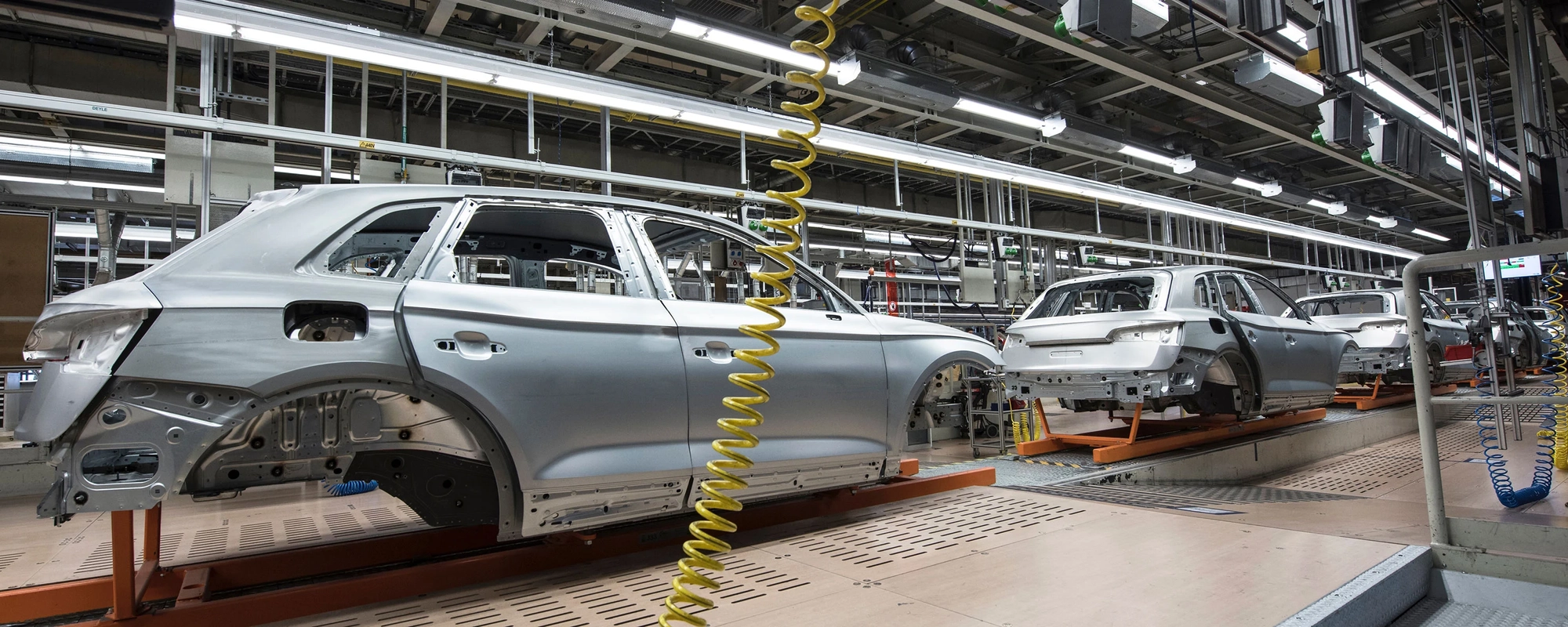 A car manufacturing assembly line with several partially assembled silver car bodies on conveyor belts. The factory has overhead lighting, yellow coiled air hoses hanging from the ceiling, and various machinery and equipment used in the assembly process.