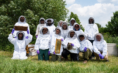 A large group of about fifteen people wearing white beekeeping suits with mesh hoods and purple gloves. Some are standing, and some are kneeling in the grassy foreground. Several people are holding frames of honeycomb or beekeeping equipment. Trees and green foliage are in the background under a blue sky with some clouds.