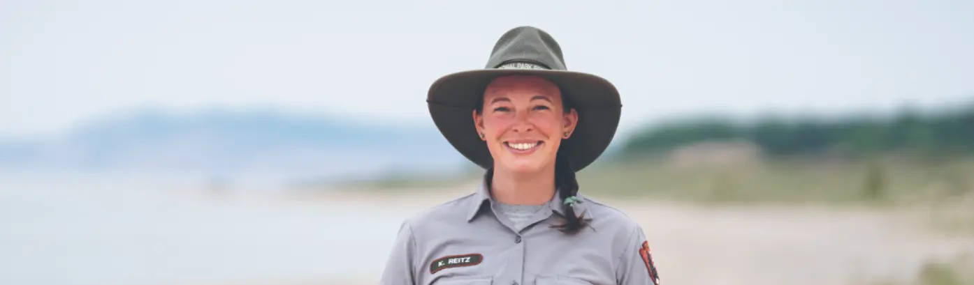 A smiling National Park Service ranger in uniform and sun hat stands near the water at Sleeping Bear Dunes.
