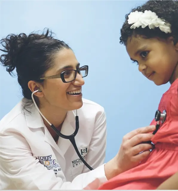 A pediatrician smiles while listening to a young girl's heartbeat with a stethoscope during a checkup.