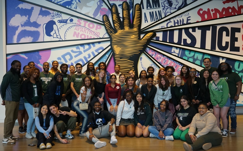 A photo of a group of students of mixed races and genders standing in front of a large mural of a hand and word art behind it.