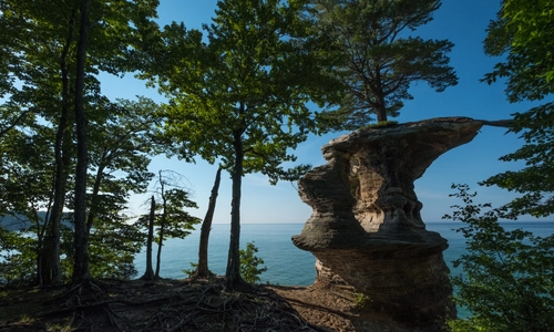 A rock formation surrounded by trees with a lake and blue sky in the background.
