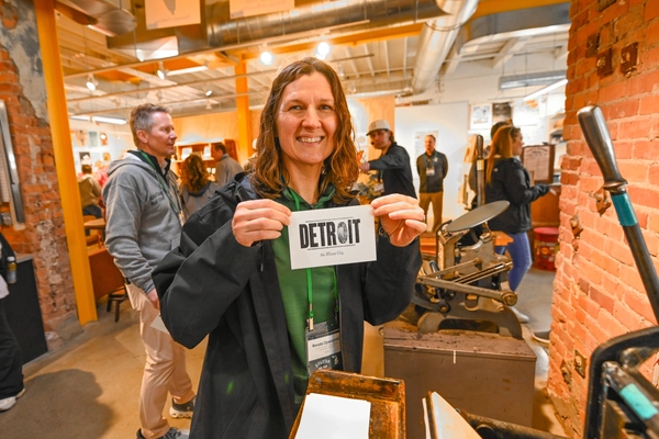 Woman holds a piece of paper saying Detroit while standing in a brick building.